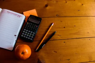 An open dated notebook, calculator, pencil and pen sit on a wooden table.