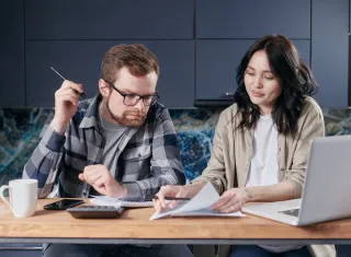 A couple reviews papers together at a kitchen table with a calculator, notebook and laptop.