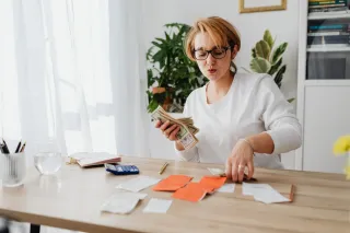 A woman sorts cash and orange and white receipts at a home table with a calculator and notebook.