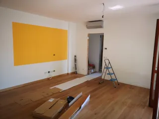An unfinished apartment room has a stepladder, exposed ceiling wiring, unpacked materials and protective floor coverings.