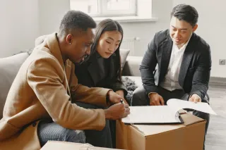 A couple reviews and signs documents with another person while seated beside moving boxes in a living room.