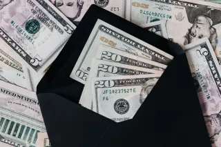US dollar banknotes sit inside an open black envelope with more notes spread underneath.
