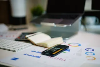 A smartphone calculator sits on printed charts beside a laptop.