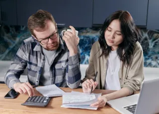 A couple uses a calculator and writes on household paperwork at their kitchen table beside a laptop.