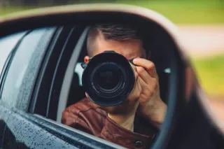 A photographer holds a camera beside a car window, visible in the side mirror.
