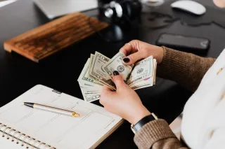 Hands count dollar-denominated banknotes beside an open planner and pen.