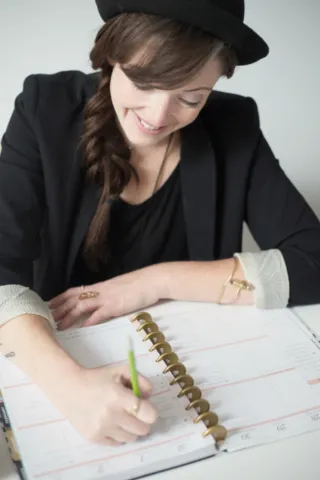 A person writes with a green pencil in an open ring-bound weekly planner.