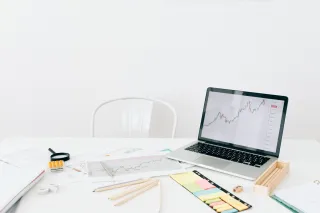 A laptop displaying market charts sits beside paperwork and stationery.