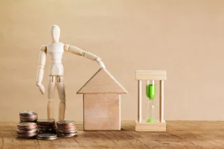 A small house model, coins, an hourglass and a wooden figure arranged on a tabletop.