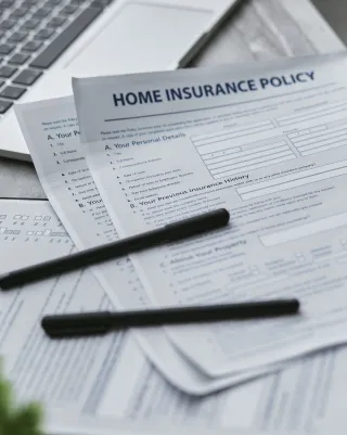 Home insurance policy forms and two black pens lie beside a laptop on a desk.