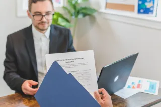 Hands hold a residential mortgage application and blue folder across a desk from another person.