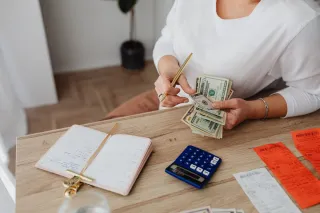 A woman counts US dollar bills beside receipts, a blue calculator and an open dated planner on a wooden desk.