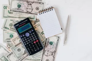 A scientific calculator and small lined notebook sit beside a pen over dollar banknotes.