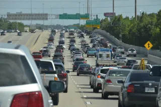 Cars and pickup trucks queue on a multilane highway beneath road signs in daylight.