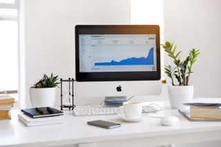 Desktop monitor displaying an analytics dashboard with a blue area chart, beside a keyboard, notebook, phone and potted plants.