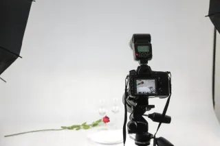 A camera on a tripod and studio lights face a tabletop arrangement of glassware and a rose.