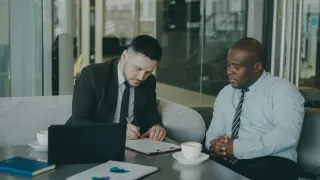 Two men seated at a table, one signing a document on a clipboard beside a laptop, printed charts and coffee cups.