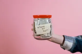 A hand holds a glass jar containing dollar banknotes and a handwritten Where to next? label against a pink background.