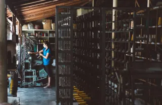 A woman operating textile machinery in a workshop lined with racks of thread spools.
