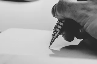 Close-up of a hand using a mechanical pencil on dotted paper, photographed in black and white.